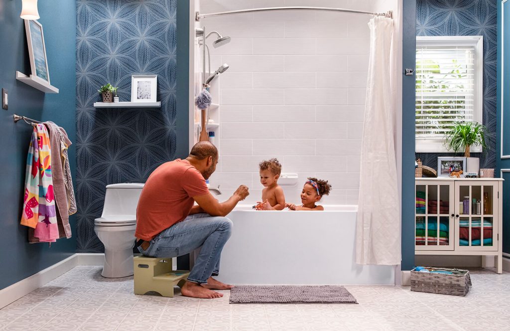 A father sits on a stool overlooking his two children in the bathtub